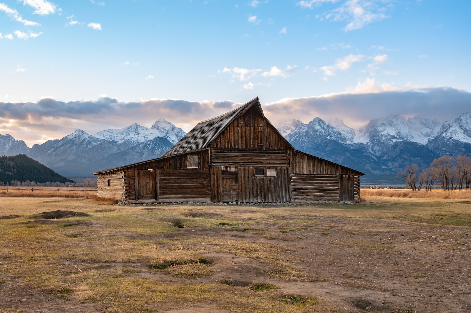brown wooden barn on brown field near snow covered mountains under blue sky during daytime